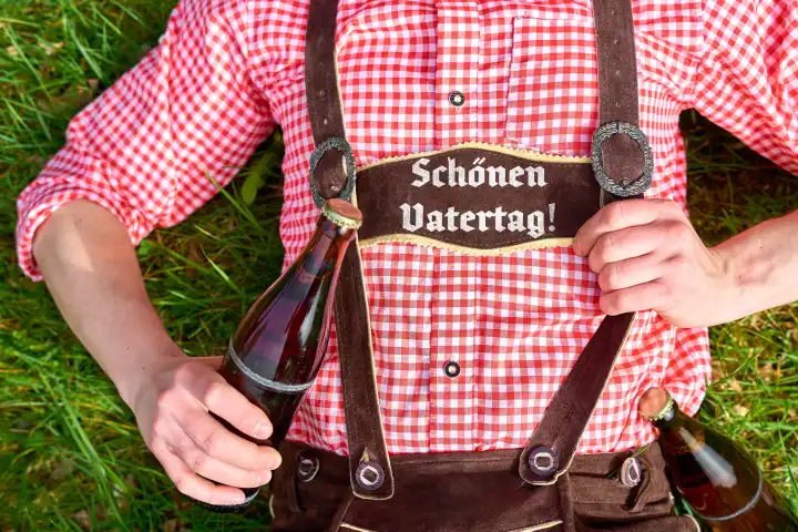 7 May 2024: Man in traditional Bavarian costume lying in a meadow with a beer bottle. Lettering on the lederhosen: Happy Father's Day! PHOTOMONTAGE