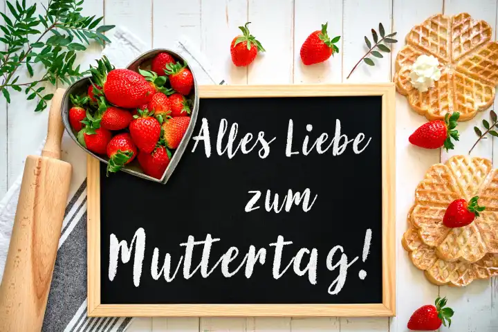 Alles Liebe zum Muttertag! Schriftzug auf einer Tafel neben frischen Erdbeeren und gebackenen Waffeln. FOTOMONTAGE