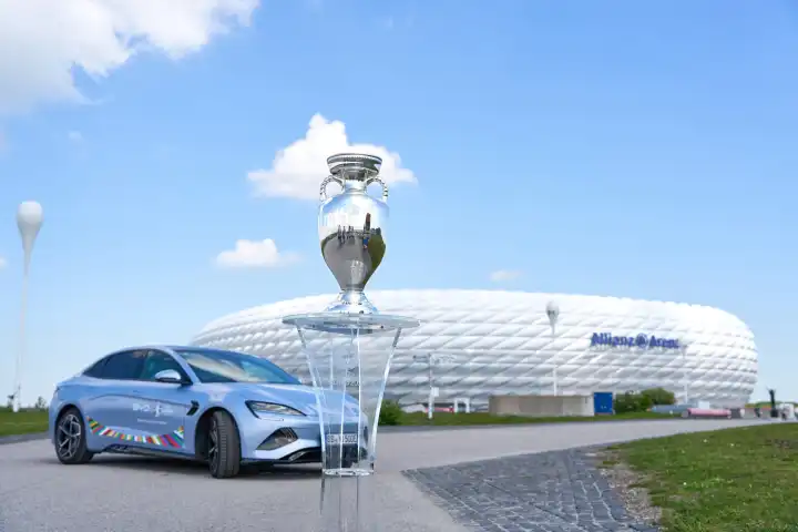BYD E-Auto mit UEFA Logo vor dem Fußball Stadion der Allianz Arena in München zusammen mit dem Siegerpokal, Henri-Delaunay-Pokal . Offizieller E-Mobilitätspartner der UEFA Euro 2024