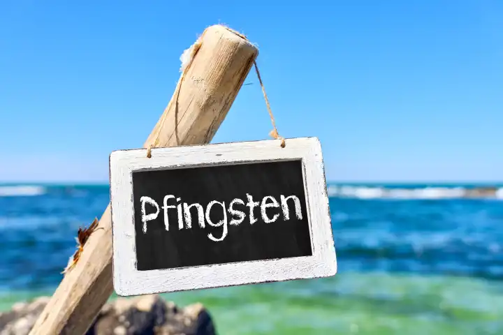 Schild aus Holz am Meer bei Sonnenschein mit der Aufschrift: Pfingsten. Pfingst-Feiertage Symbolbild. FOTOMONTAGE