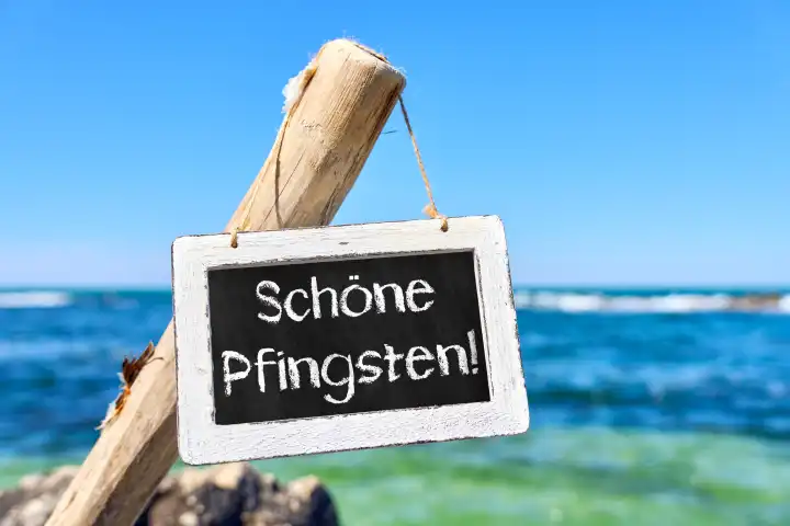 Schild aus Holz am Meer bei Sonnenschein mit der Aufschrift: Schöne Pfingsten. Pfingsten, Feiertag Symbolbild. FOTOMONTAGE