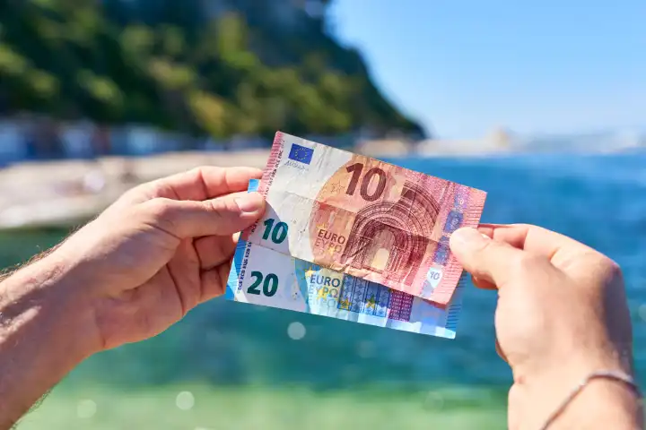 Ein Mann hält Euro Geldscheine vor das Meer am Strand von Ancona in Italien. Urlaubsgeld und Urlaubsnebenkosten Konzept