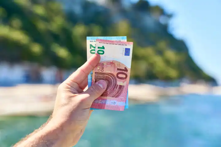 Ein Mann hält Euro Geldscheine vor das Meer am Strand von Ancona in Italien. Urlaubsgeld und Urlaubsnebenkosten Konzept