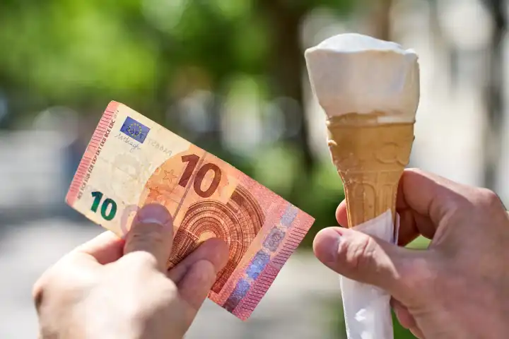 Hand hält ein Eis in einer Waffel neben einen 10 Euro Bargeld Geldschein. Symbolbild Kosten und erhöhung für den Preis einer Kugel Eis