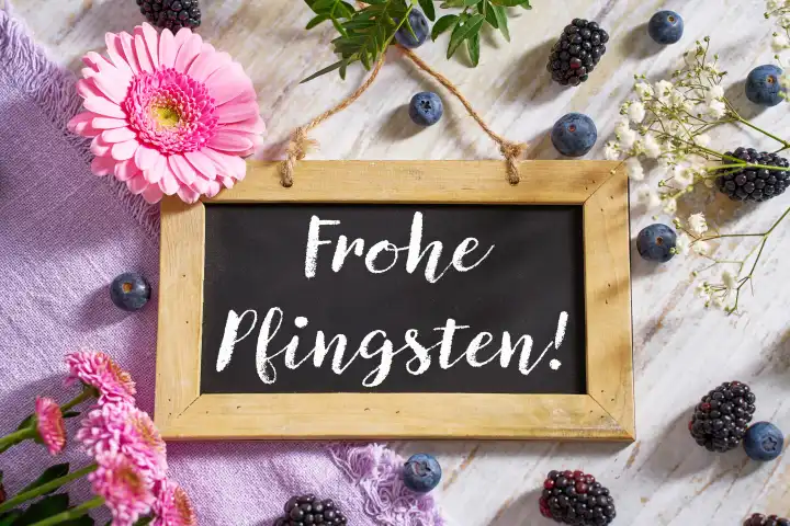 Schild mit Aufschrift: Frohe Pfingsten! umgeben von bunten Blumen und Dekoration. Pfingsten. FOTOMONTAGE