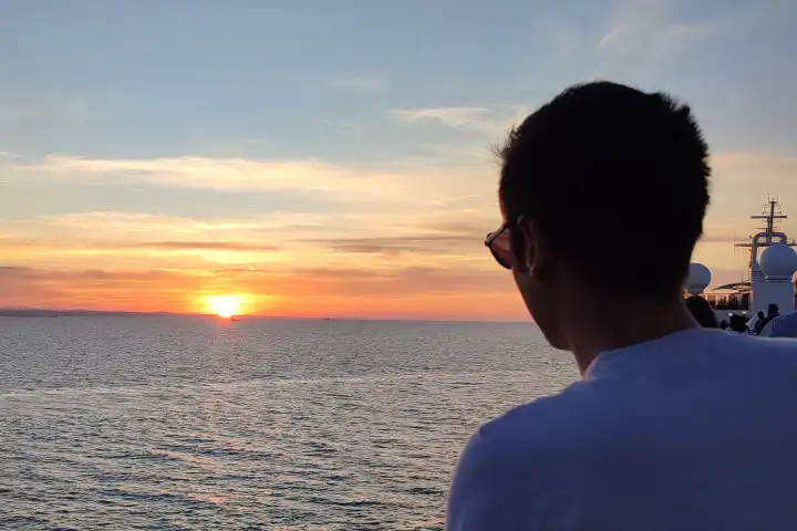 Mann mit Sonnenbrille blickt von einem Kreuzfahrtschiff in den Sonnenuntergang auf das weite Meer im Sommerurlaub