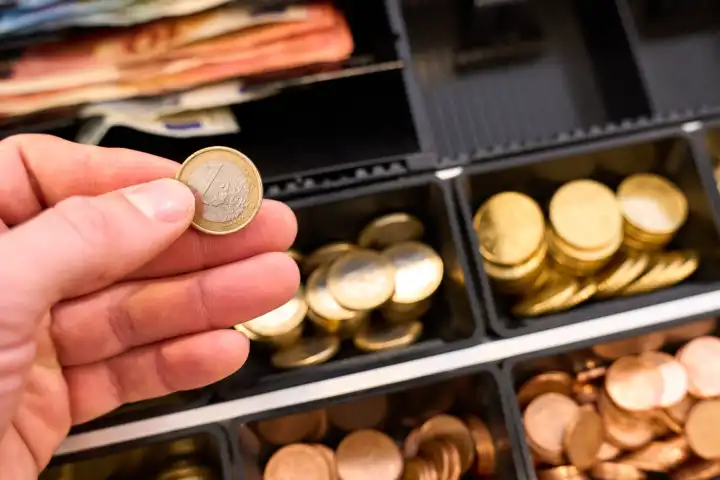 Hand hält eine 1 Euro Münze vor eine Kasse in einem Geschäft