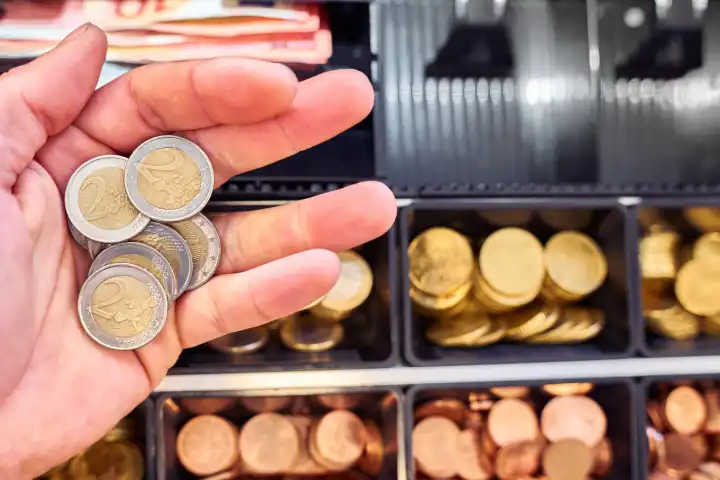Hand hält zwei Euro Wechselgeld Bargeld Münzen vor eine Kasse in einem Geschäft