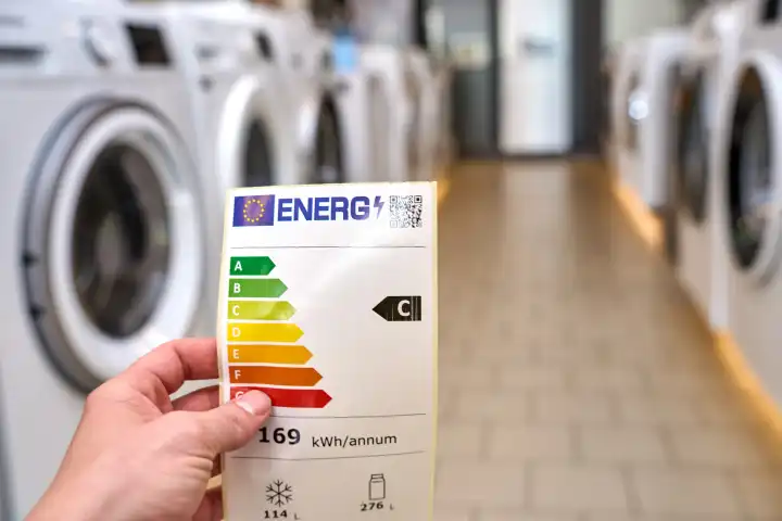 Eine Hand hält ein Energie Label der Europäischen Union in einem Geschäft mit Haushaltsgeräten wie Waschmaschinen