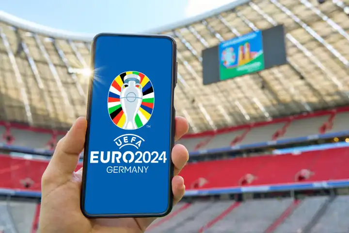 Hand hält in der Allianz Arena in München ein Smartphone mit dem UEFA EURO 2024 Logo. Symbolfoto Fußball Europameisterschaft in Deutschland. FOTOMONTAGE