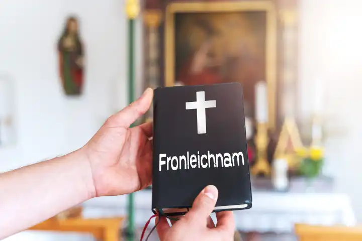 Hand hält in einer Kirche ein schwarzes Buch mit Aufschrift: Fronleichnam. FOTOMONTAGE