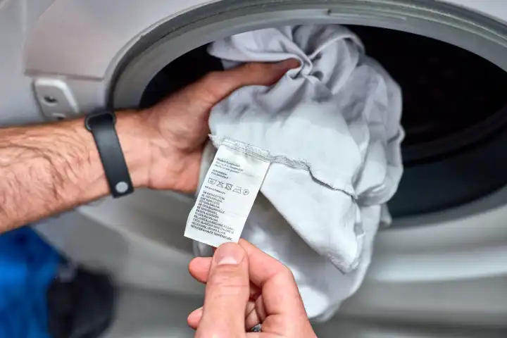                                Germany, Bavaria 21 May 2024: Man looks at the label of a garment or T-shirt of his clothes in front of the washing machine and checks in which program and at which temperature it has to be washed - washing instructions