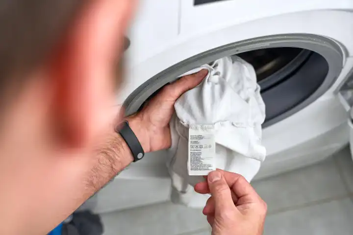 Mann schaut sich vor der Waschmaschine das Etikett eines Kleidungsstückes, bzw. T-Shirt seiner Kleidung an und prüft in welchem Programm und auf welcher Temperatur es waschen muss - Waschanleitung