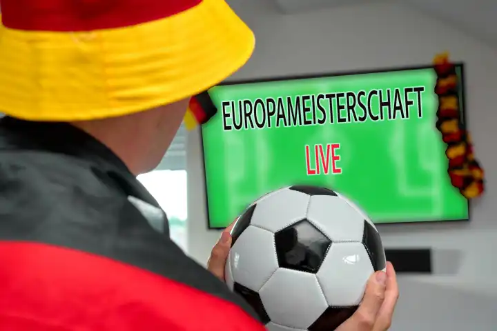 Fußballfan in Deutschlandfarben hält gespannt einen Fußball und verfolgt die Live-Übertragung der Europameisterschaft auf einem Fernseher Zuhause. FOTOMONTAGE