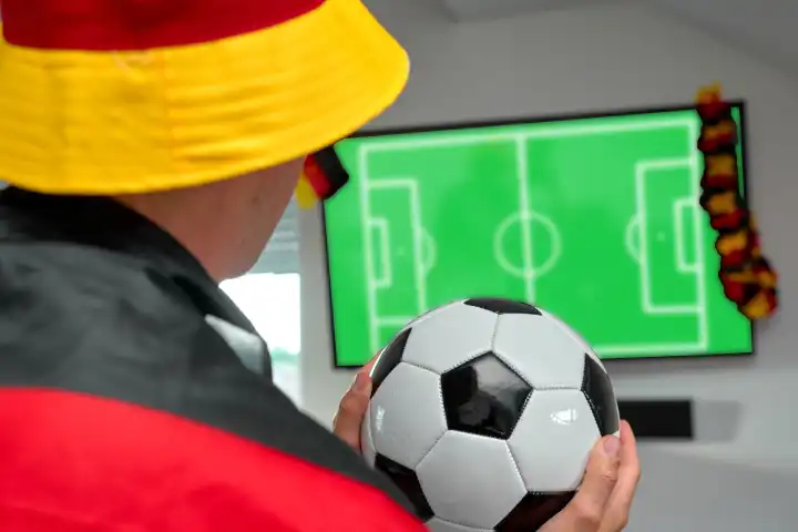 Fußballfan in einem Deutschlandhut jubelt einem Tor zu, während er das Spiel der Europameisterschaft live auf seinem Heimfernseher verfolgt, eingehüllt in eine Deutschlandflagge, symbolisch für nationale Unterstützung und Sportbegeisterung. FOTOMONTAGE