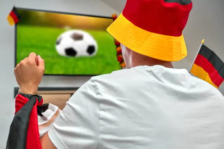 Fußballfan in einem Deutschlandhut jubelt einem Tor zu, während er das Spiel der Europameisterschaft live auf seinem Heimfernseher verfolgt, eingehüllt in eine Deutschlandflagge, symbolisch für nationale Unterstützung und Sportbegeisterung. FOTOMONTAGE