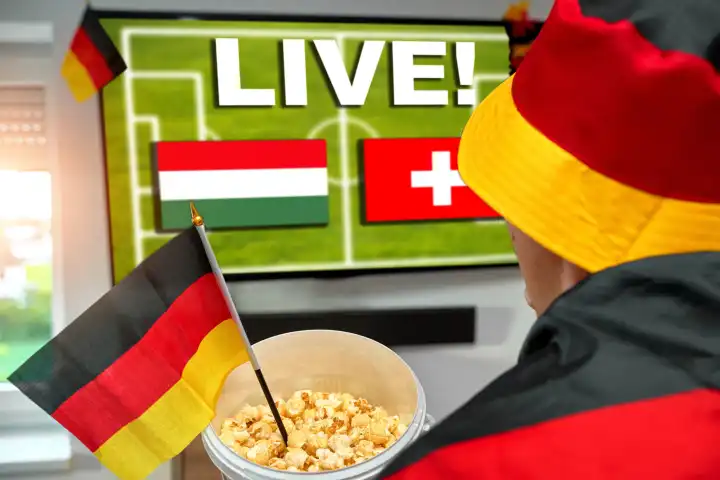 Ein deutscher Fußballfan schaut das Fußball Europameisterschaft Spiel zwischen Ungarn und der Schweiz live im Fernsehen. Mit Deutschland-Fanartikel, Popcorn und der deutschen Flagge genießt er das spannende Event von zu Hause im Wohnzimmer. FOTOMONTAGE