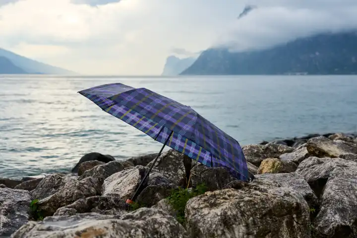  Nago Torbole, Lake Garda, Italy - 25 June 2024: Symbolic image of thunderstorms and rain on vacation at Lake Garda. An umbrella lies on the northern shore in front of the lake under a cloudy sky