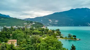 Riva del Garda, Lake Garad, Italy - June 25, 2024: Shows the picturesque Lake Garda, surrounded by lush green forests and high mountains that provide an impressive backdrop. Charming buildings and well-tended green areas can be seen on the shore, inviting you to take a stroll and relax.