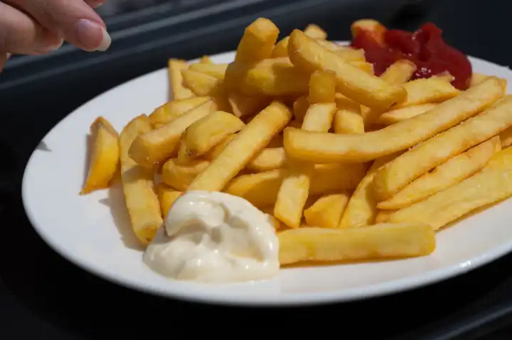 Hand of woman taking fresh French fries with ketchup and mayonnaise on white plate in sunlight