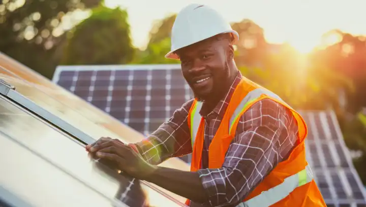 dark skinned man wearing an orange safety vest and works with solar panel, sun shines in background