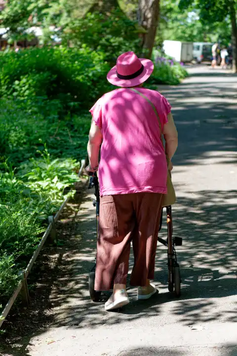 An older woman in a pink blouse and a pink hat walks through a park