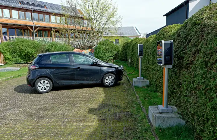 Ein E-Auto wird an einer freistehenden  Ladesäule auf einem Parkplatz vor einer Hecke geladen