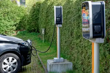 An electric car is charged at a free-standing charging station in a parking lot in front of a hedge
