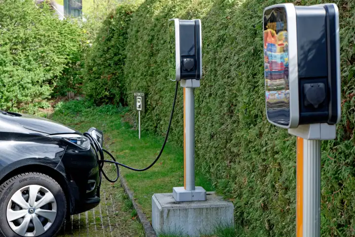 An electric car is charged at a free-standing charging station in a parking lot in front of a hedge