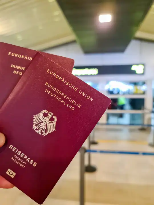A hand holds two German passports in front of a soft travel airport background on vacation