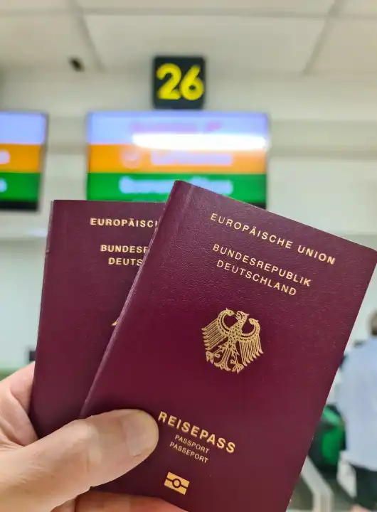 A hand holds two German passports in front of a soft travel airport background on vacation